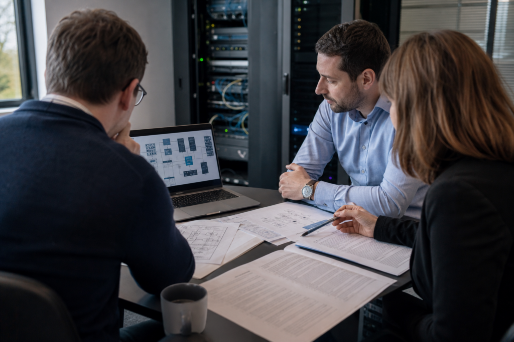 Réunion IT en PME autour d’un contrat d’infogérance avec infrastructure réseau, postes de travail et équipements critiques en arrière-plan dans un environnement professionnel moderne